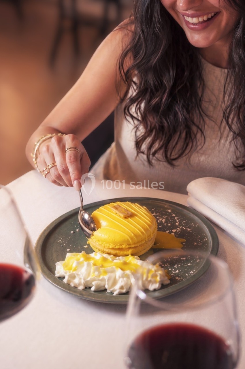 Femme souriante dégustant un dessert jaune avec de la crème fouettée, entourée de verres de vin rouge.