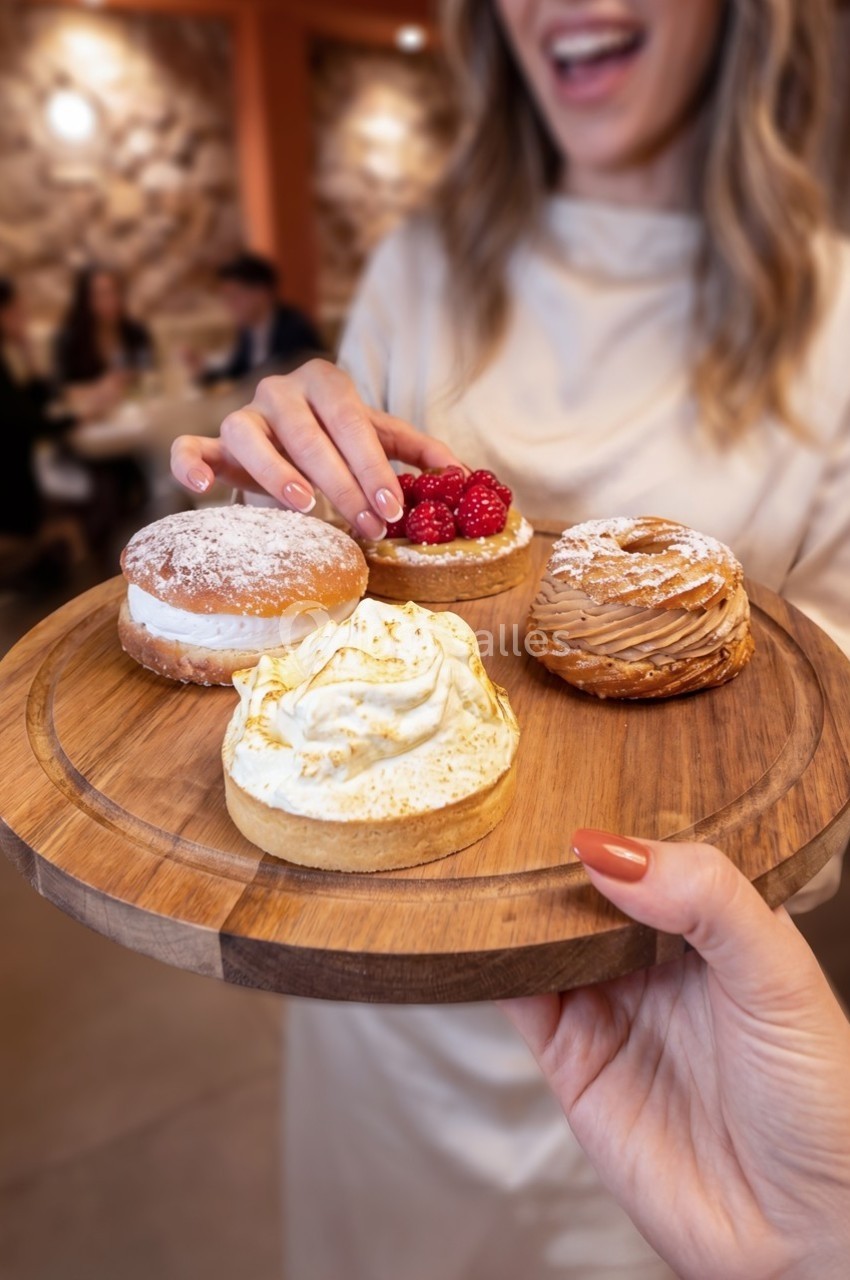 Plateau en bois avec quatre pâtisseries variées, une main en sélectionne une, en arrière-plan une femme souriante.