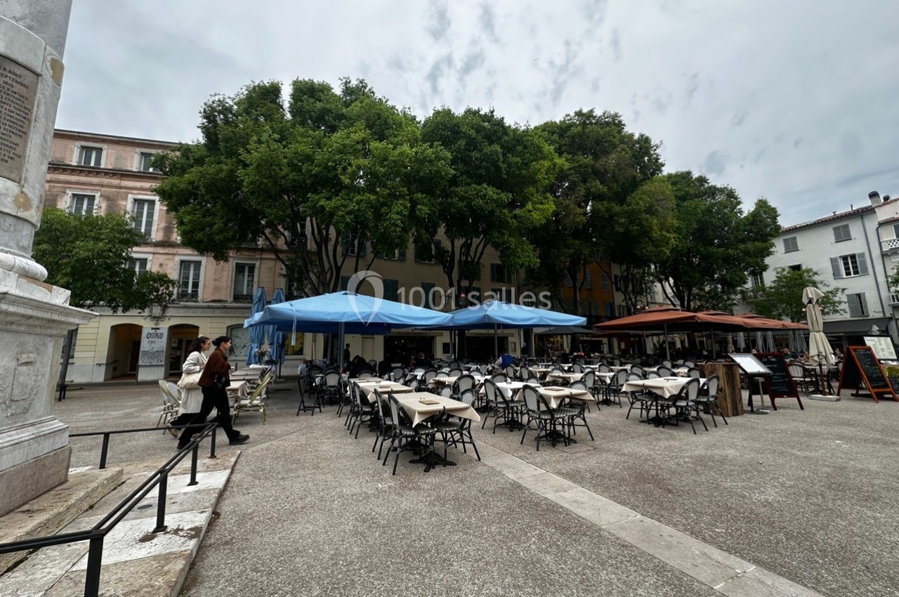 Terrasse de café avec tables et chaises vides sous des parasols bleus et rouges, entourée d'arbres et de bâtiments.