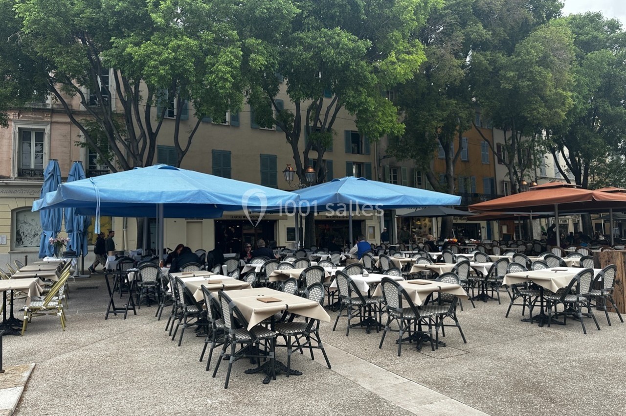 Terrasse de café avec tables et chaises vides sous des parasols bleus et orange, entourée d'arbres et de bâtiments.