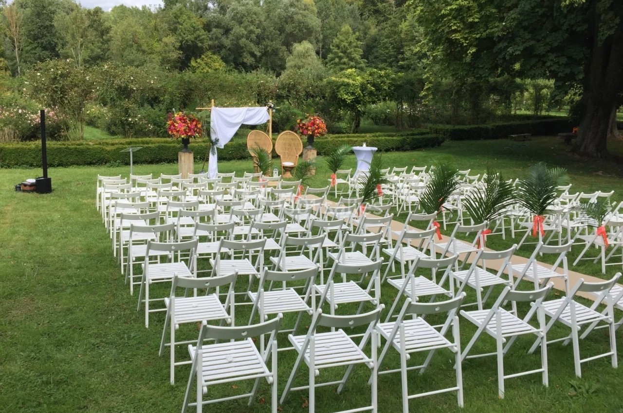 Chaises blanches alignées en extérieur devant une arche décorée pour une cérémonie, entourées de verdure.