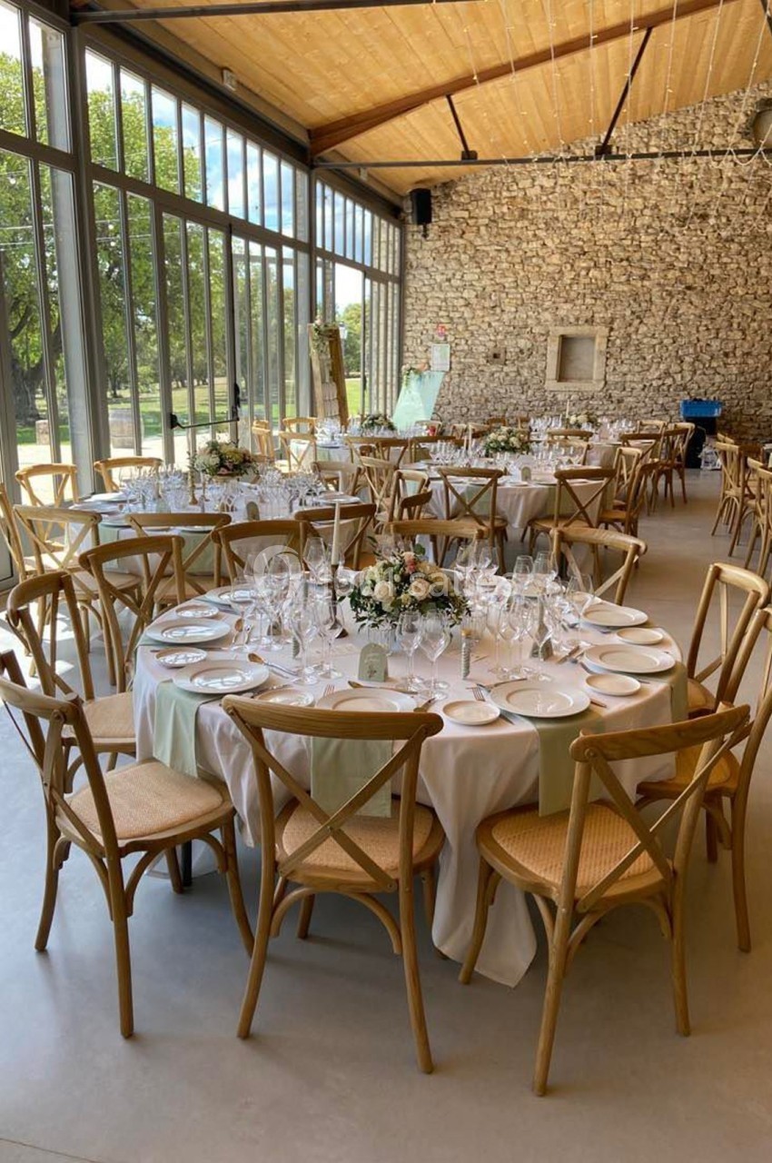 Salle lumineuse avec tables rondes dressées pour un événement, décorées de fleurs et entourées de chaises en bois.