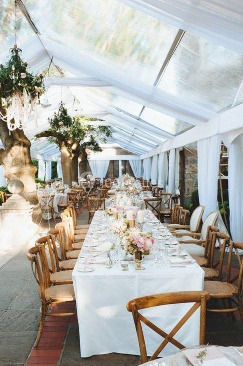 Tables de banquet décorées de fleurs sous une tente transparente avec chaises en bois et lumière naturelle.