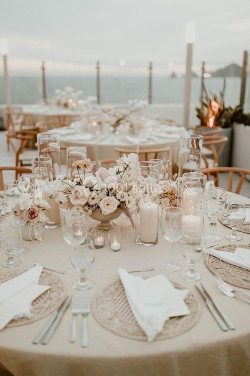 Table élégamment dressée en extérieur avec des fleurs blanches, des bougies et une vue sur la mer au coucher du soleil.