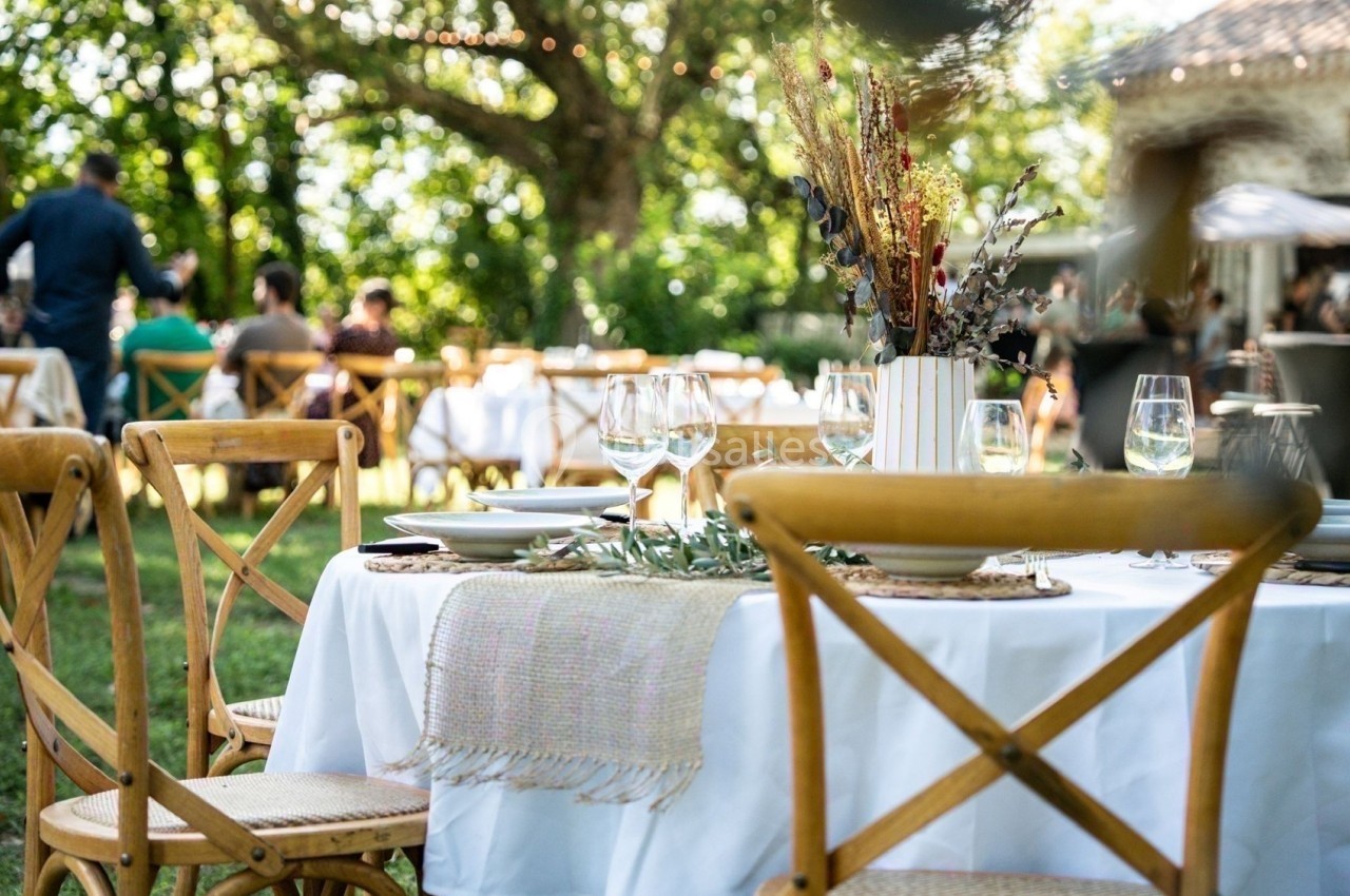 Table dressée en extérieur avec nappes blanches, verres et décorations florales, entourée de chaises en bois.
