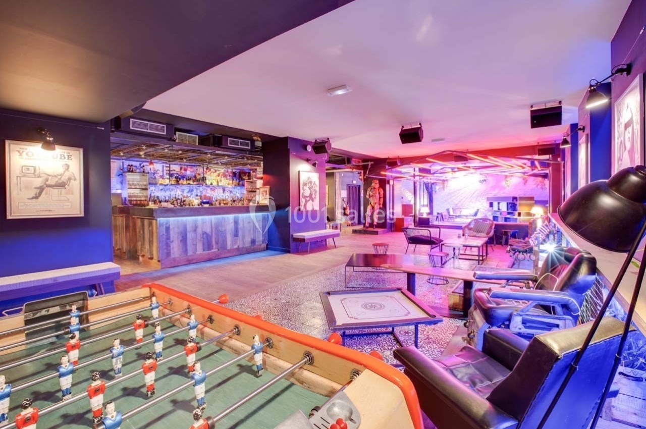 Salle de loisirs moderne avec baby-foot, bar éclairé, fauteuils et ambiance lumineuse colorée.