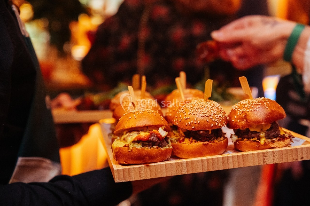 Plateaux de mini-burgers garnis, servis lors d'un événement avec des convives en arrière-plan flou.