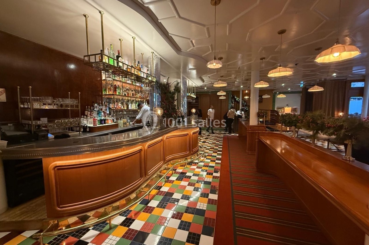 Intérieur d'un bar avec comptoir en bois, étagères de bouteilles, sol coloré et éclairage chaleureux.