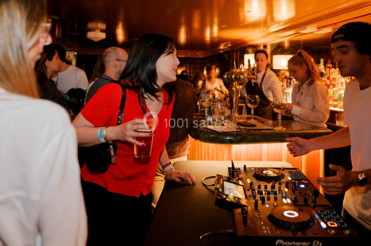 Une femme en rouge tenant un verre discute avec un DJ près de sa console dans un bar animé.