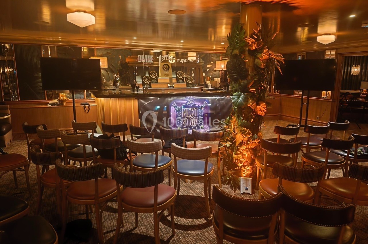 Salle aménagée avec des chaises disposées face à un bar éclairé, décoré d'un panneau lumineux et de plantes.