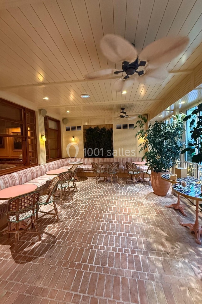 Salle lumineuse avec sol en briques, banquettes, tables, chaises en osier, plantes en pot et ventilateurs au plafond.
