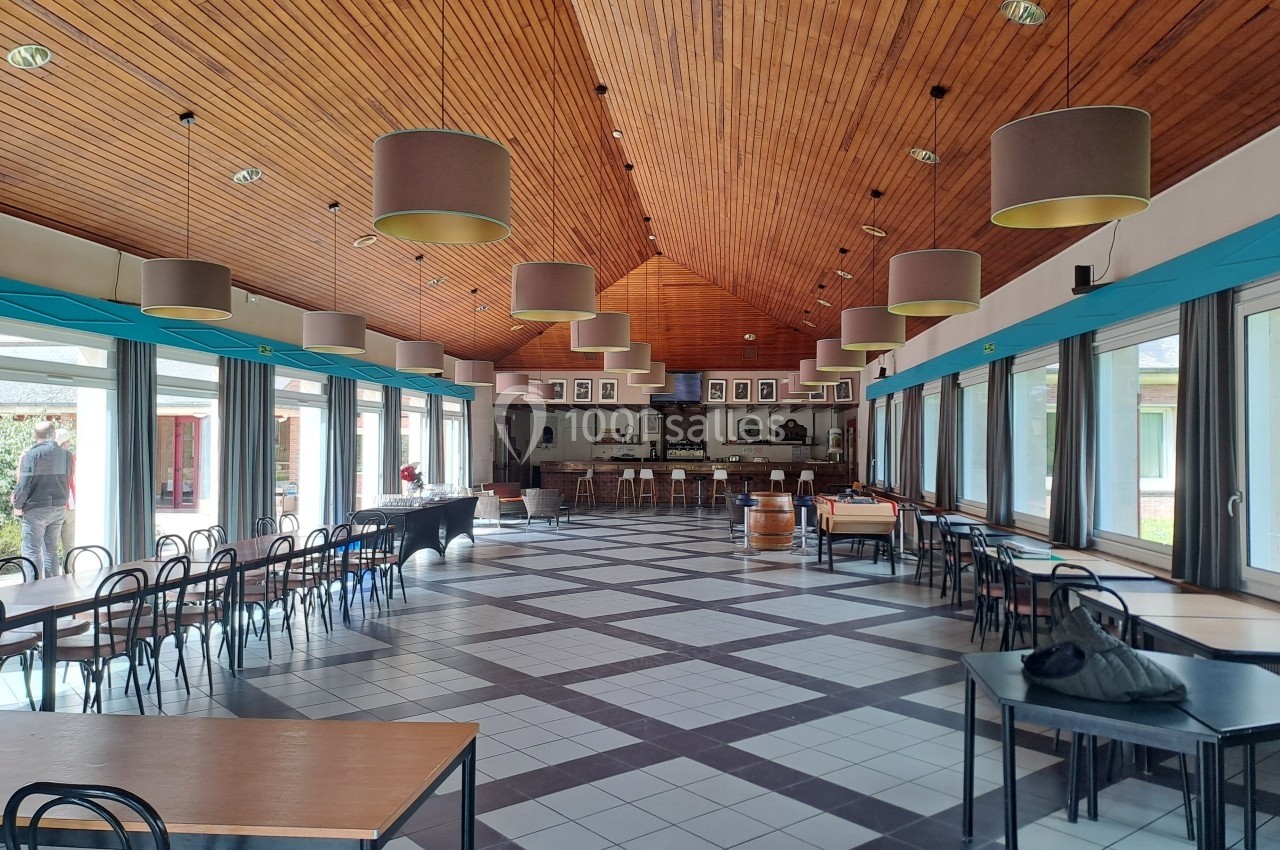 Salle spacieuse avec plafond en bois, grandes baies vitrées, tables et chaises disposées dans un espace lumineux.