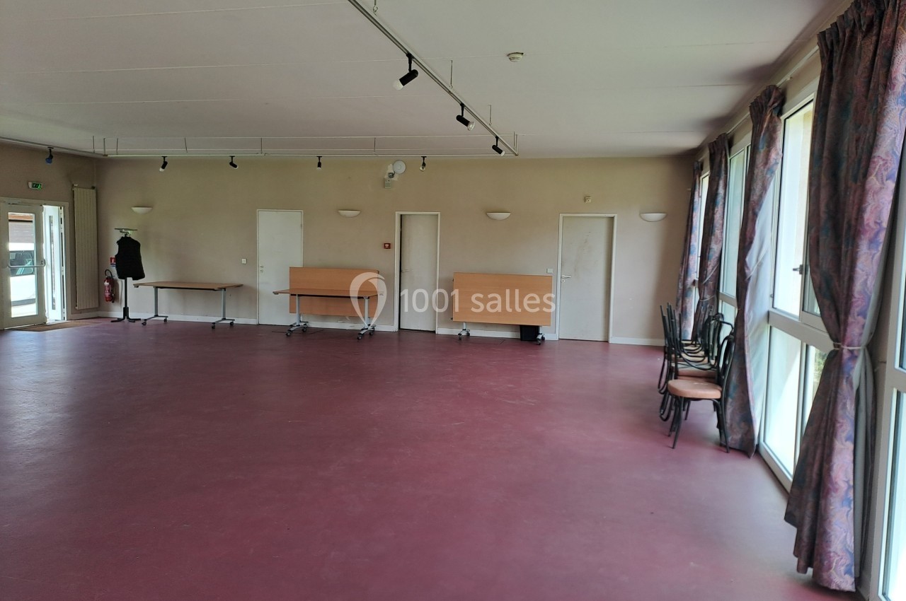 Salle vide avec sol rouge, murs clairs, grandes fenêtres avec rideaux et quelques tables et chaises empilées.