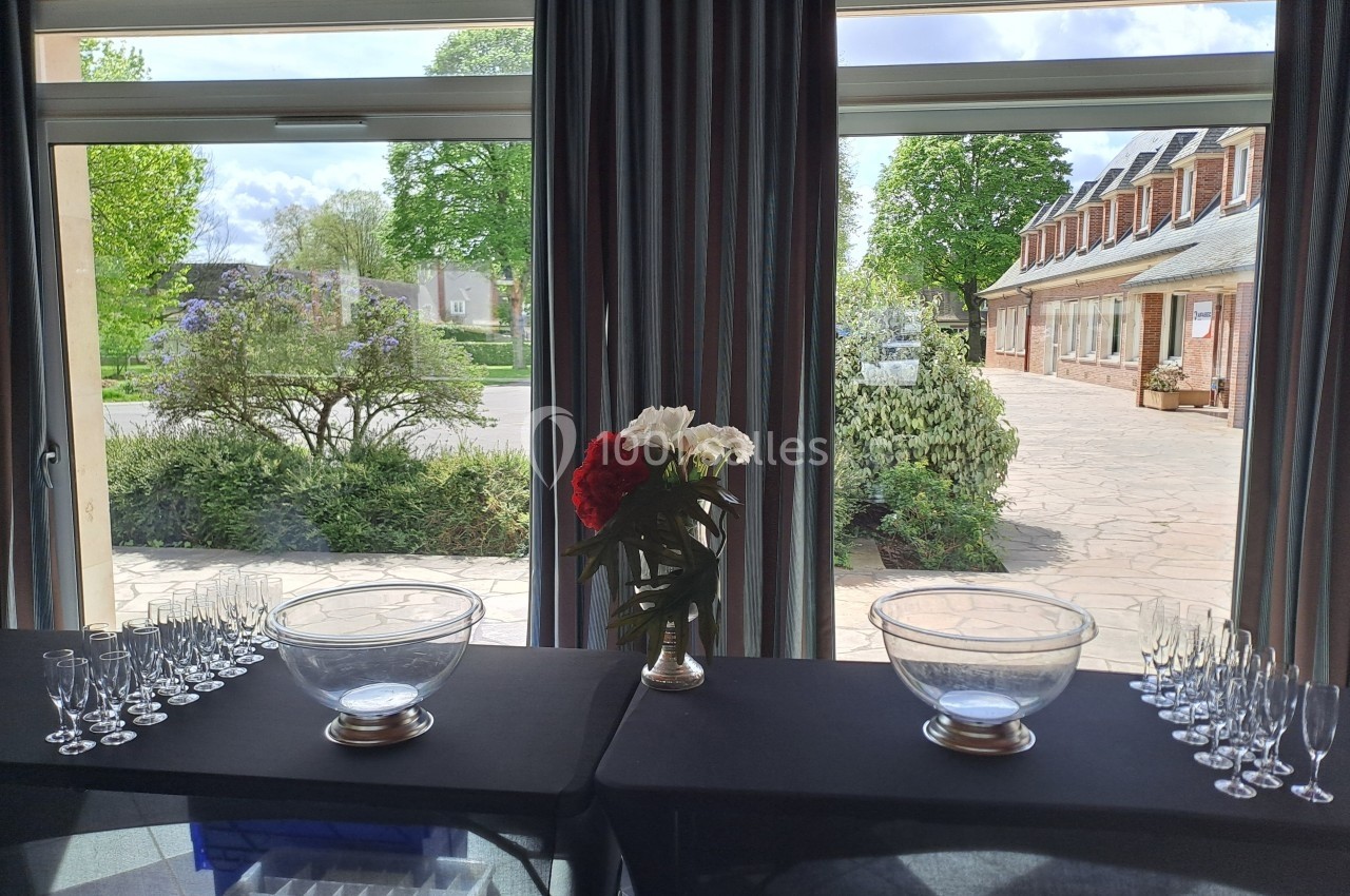 Table dressée avec des coupes et des bols en verre devant de grandes baies vitrées donnant sur un jardin et une cour pavée.