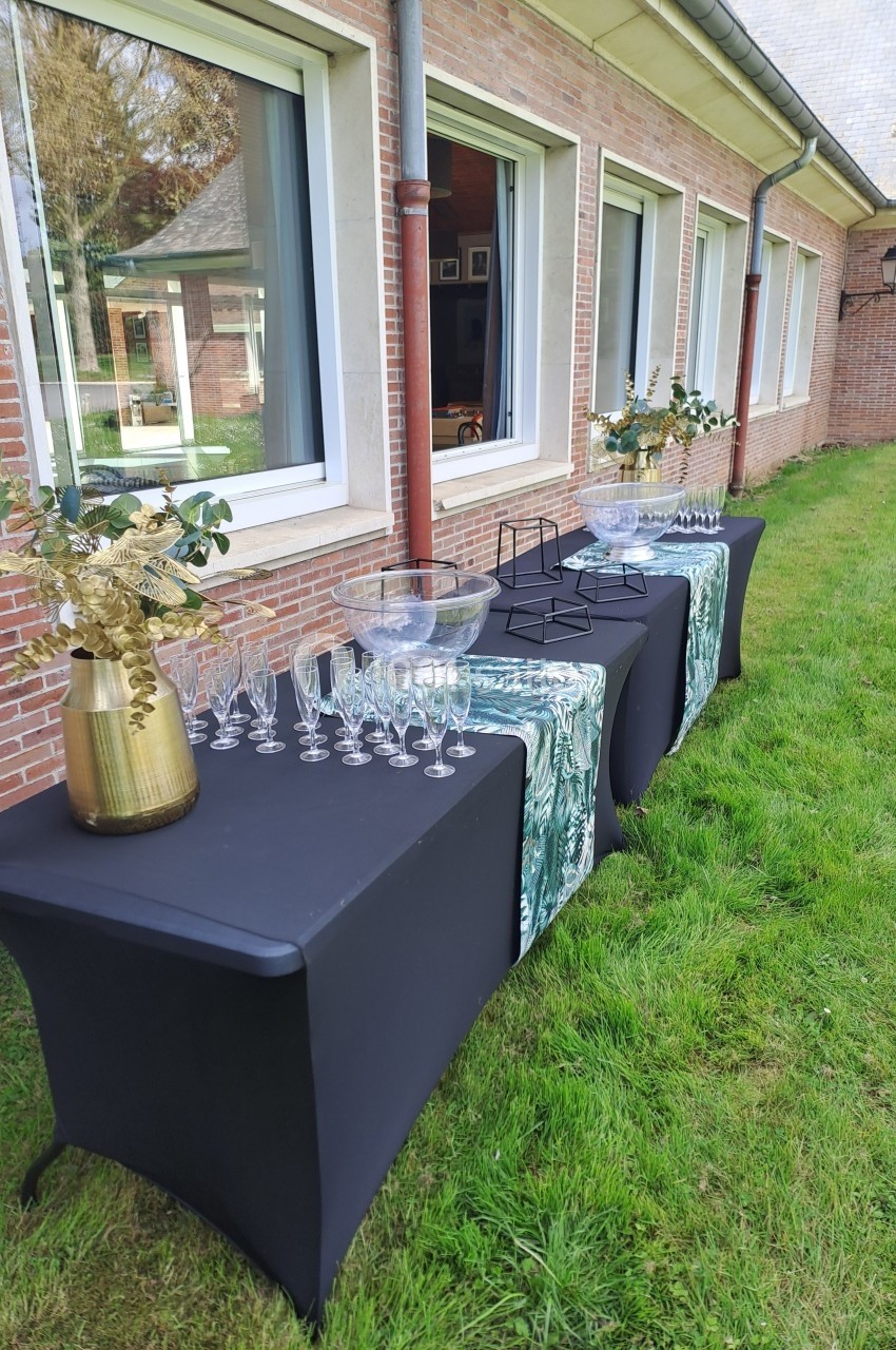 Tables noires dressées en extérieur avec verres, carafes et décorations florales devant un bâtiment en briques.