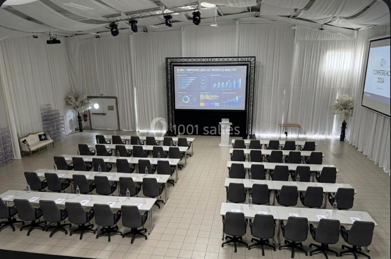 Salle de conférence aménagée avec des rangées de tables et chaises, écran de projection et pupitre au centre.