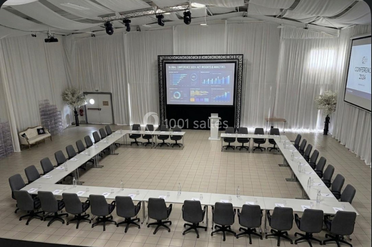 Salle de conférence avec disposition en U, tables blanches, chaises grises et écrans affichant des graphiques.