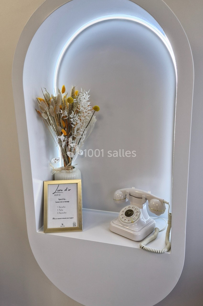 Niche murale blanche avec un téléphone rétro, un bouquet de fleurs séchées et un cadre doré contenant un texte.