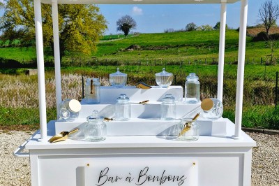 Chariot blanc avec bocaux en verre et cuillères dorées, installé en extérieur devant un paysage champêtre.