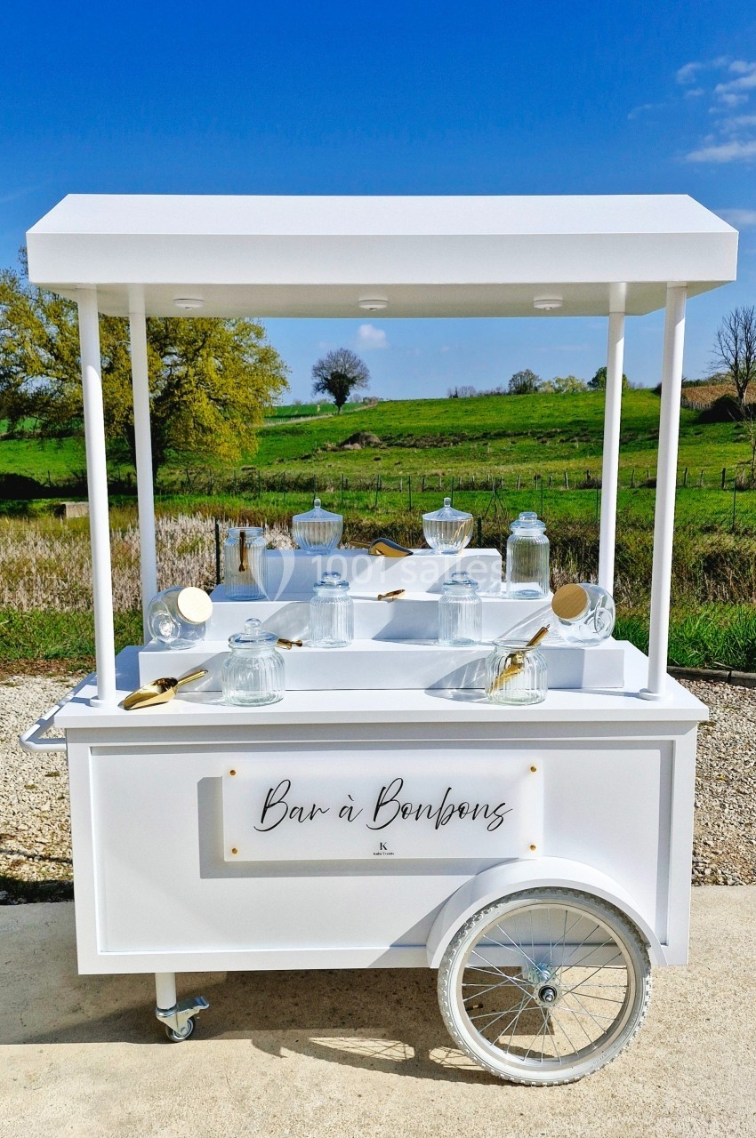 Chariot blanc avec bocaux en verre et cuillères dorées, installé en extérieur devant un paysage champêtre.