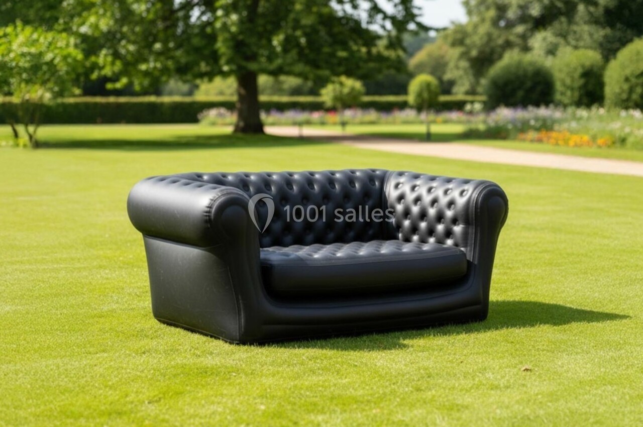 Canapé noir capitonné placé sur une pelouse verte dans un jardin avec des arbres et des parterres de fleurs en arrière-plan.
