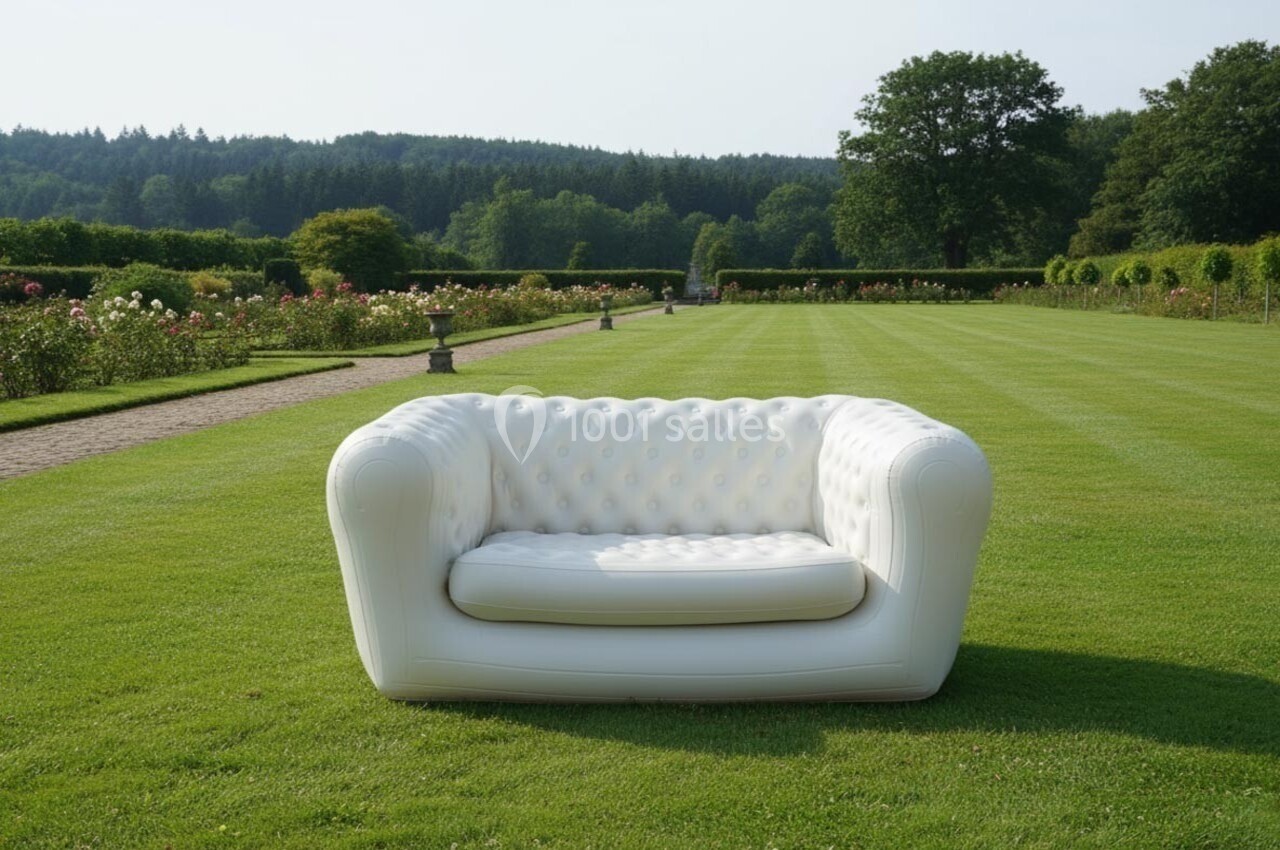 Canapé blanc capitonné placé sur une pelouse verte dans un jardin avec des haies, des fleurs et des arbres en arrière-plan.