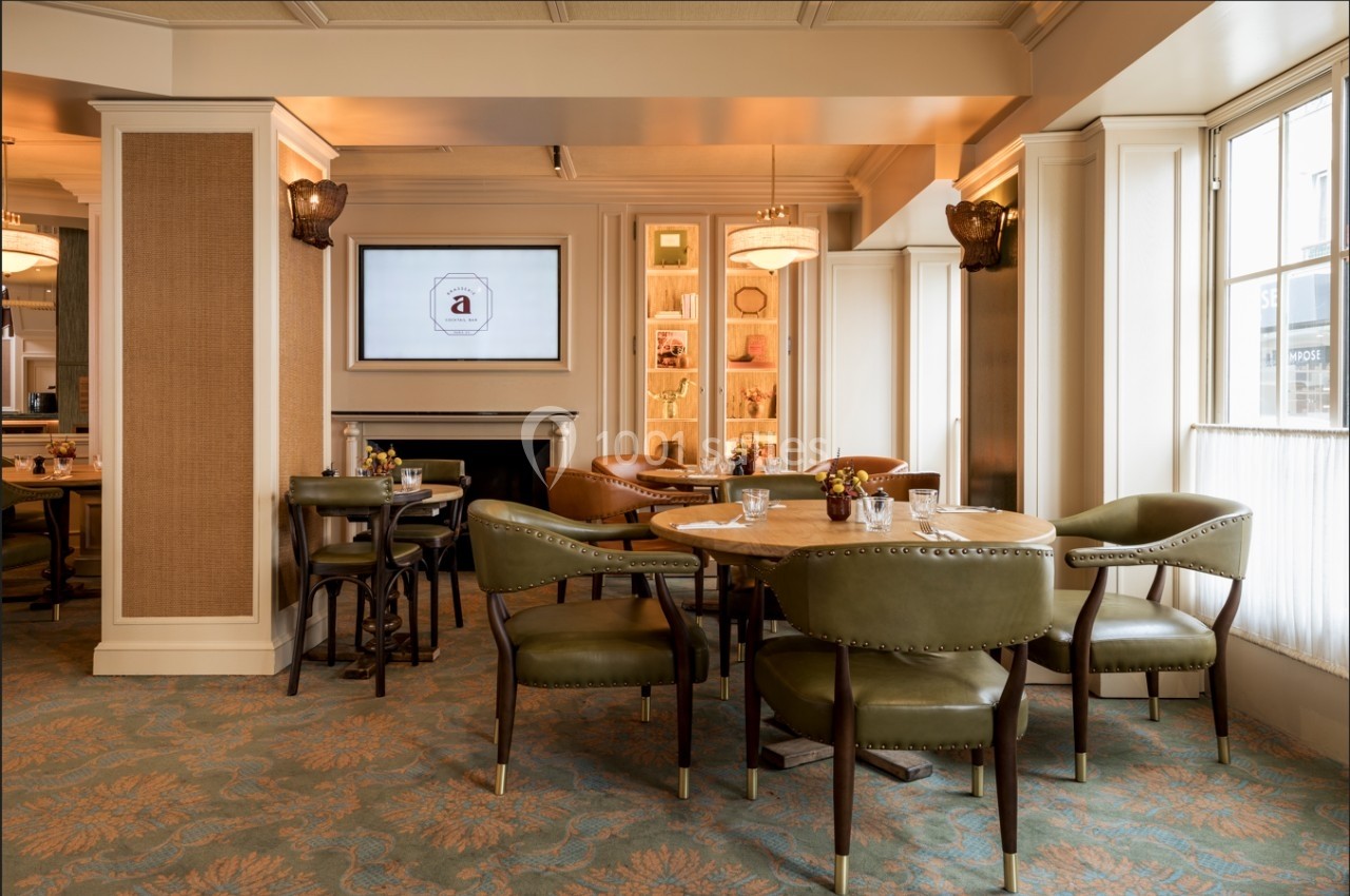 Salle de restaurant lumineuse avec tables en bois, chaises en cuir vert et écran mural allumé.