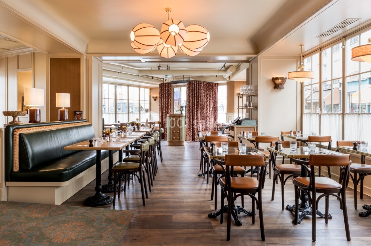 Salle de restaurant lumineuse avec tables en bois, chaises en cuir et banquette, éclairée par des suspensions design.