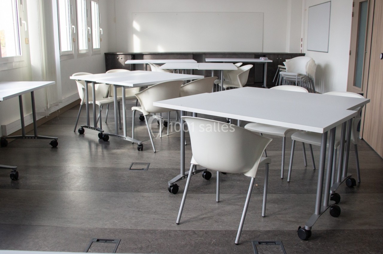 Salle de classe lumineuse avec des tables blanches sur roulettes et des chaises, entourée de fenêtres et d'un tableau blanc.