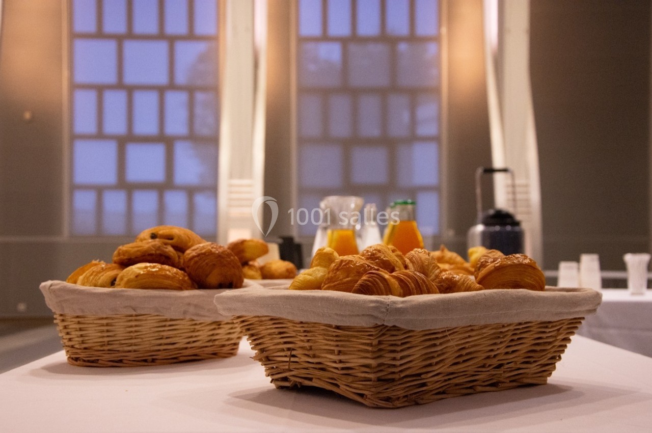 Paniers de viennoiseries avec jus d'orange et cafetière en arrière-plan dans une salle lumineuse.