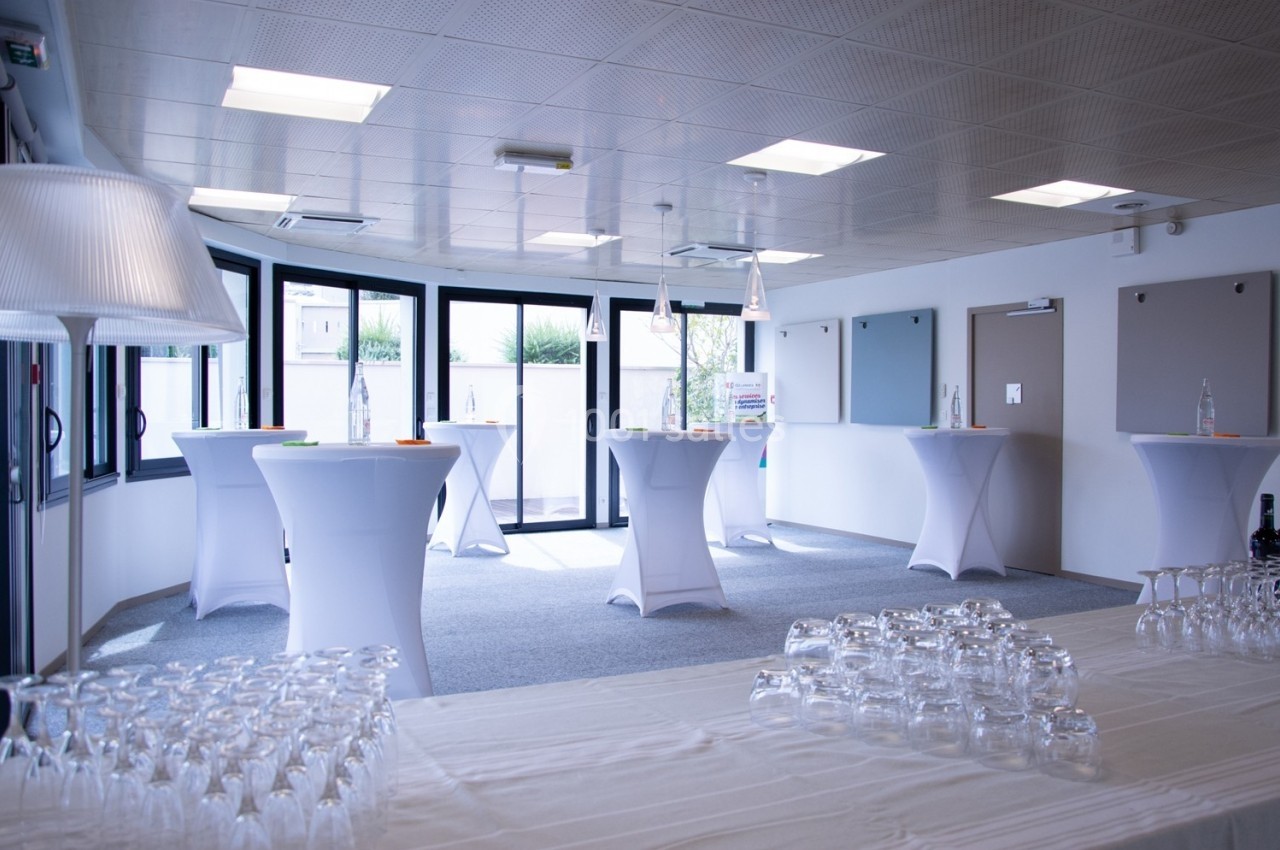 Salle lumineuse aménagée pour un événement, avec des tables hautes drapées de blanc et des verres disposés au premier plan.