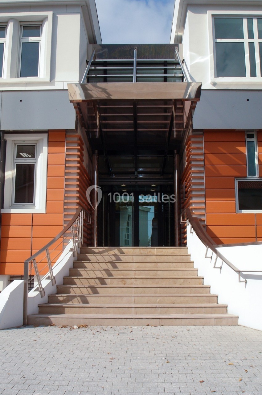 Entrée d'un bâtiment moderne avec escalier central, rampe d'accès et façade orange et blanche.
