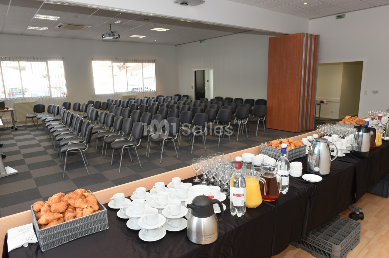 Salle de conférence avec des rangées de chaises vides et une table dressée pour un buffet petit-déjeuner.