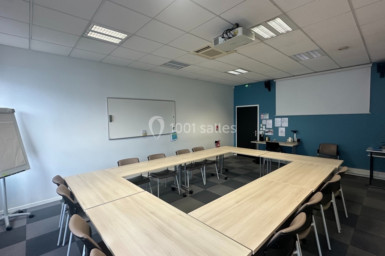Salle de réunion lumineuse avec tables disposées en U, tableau blanc, paperboard et projecteur au plafond.