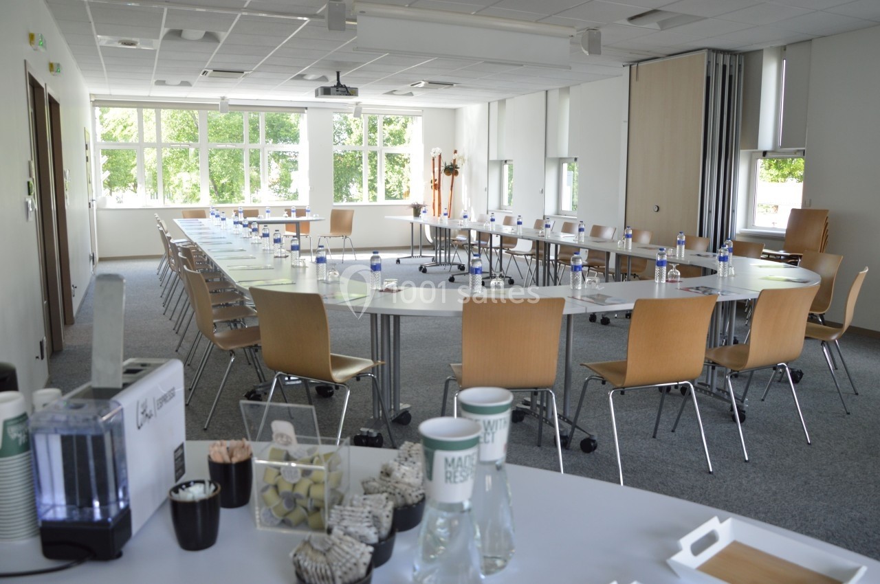 Salle de réunion lumineuse avec tables disposées en U, chaises en bois, et matériel de conférence prêt à l'usage.