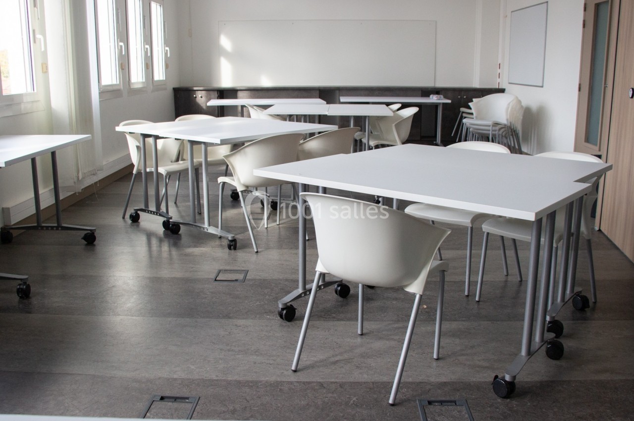 Salle de classe lumineuse avec tables blanches, chaises modernes et tableau blanc au fond.