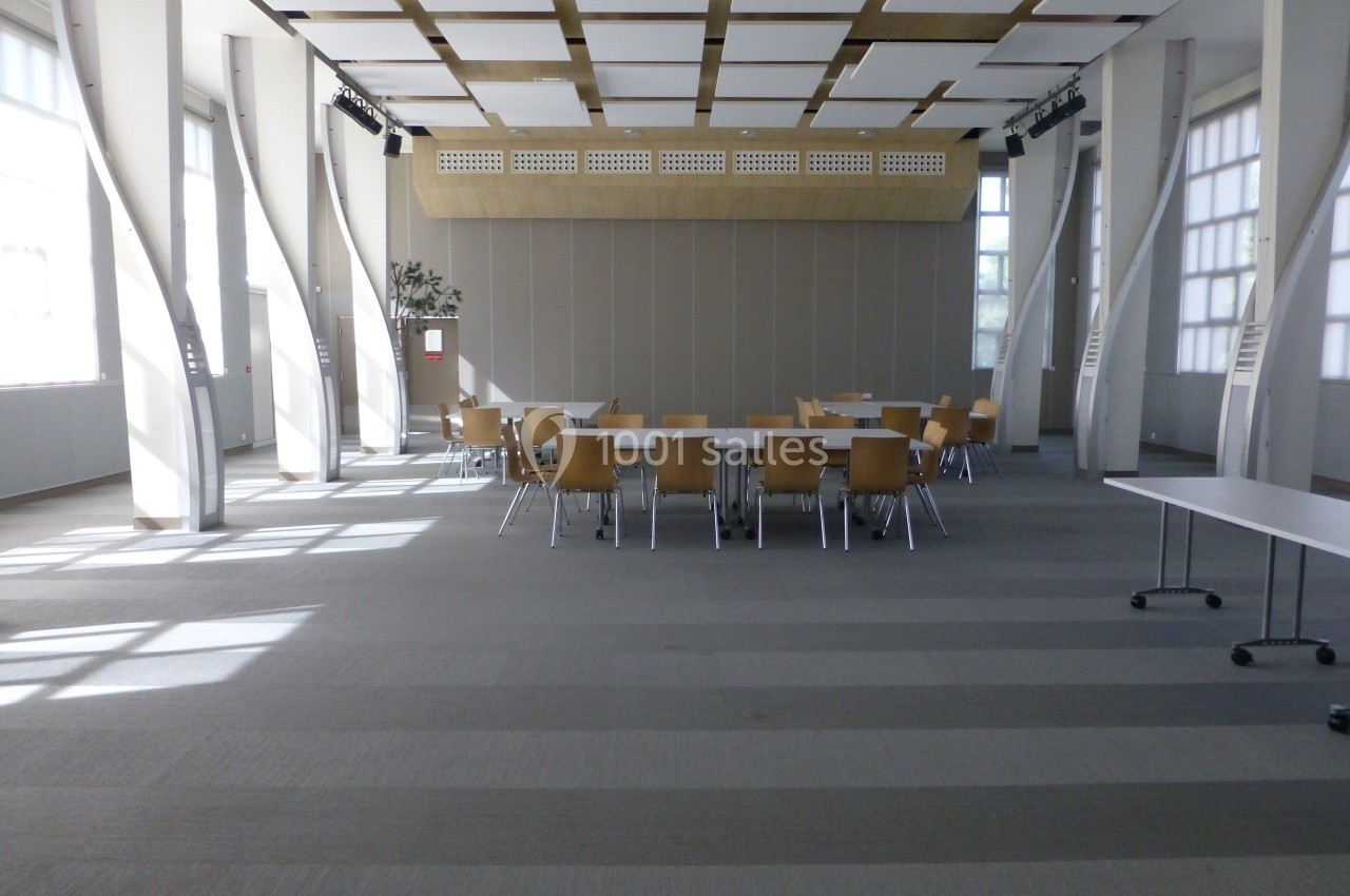 Salle de réunion lumineuse avec des tables et des chaises en bois disposées au centre, entourées de grandes fenêtres.