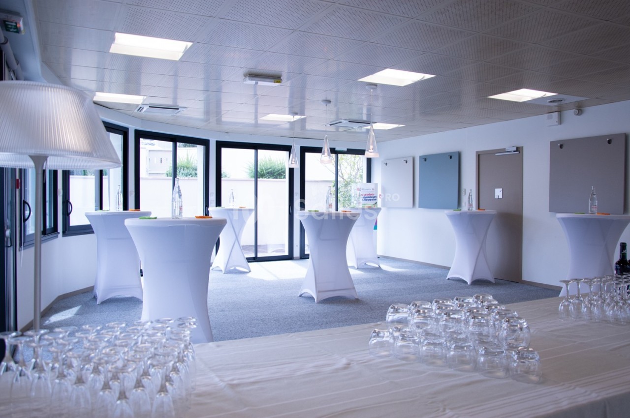 Salle lumineuse aménagée pour un événement, avec des tables hautes drapées de blanc et des verres disposés au premier plan.