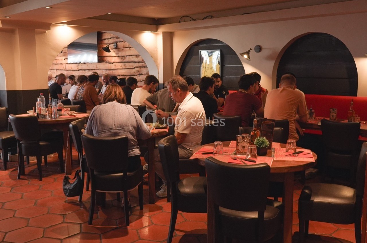 Des personnes dînent dans un restaurant à l'ambiance chaleureuse, assises à des tables rondes avec nappes rouges.