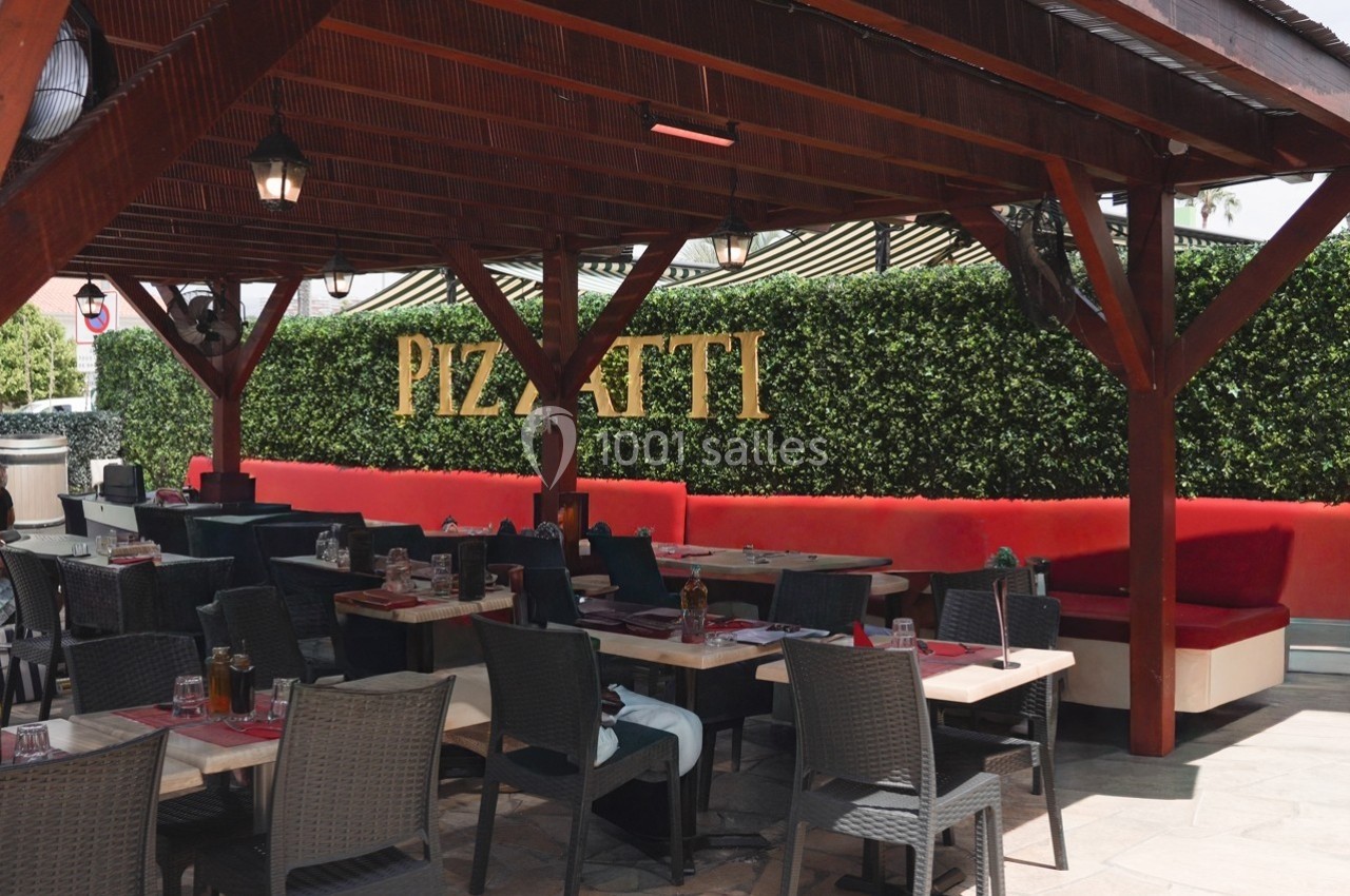 Terrasse d'un restaurant avec tables dressées, banquettes rouges et mur végétal portant l'inscription ’Pizzatti’.