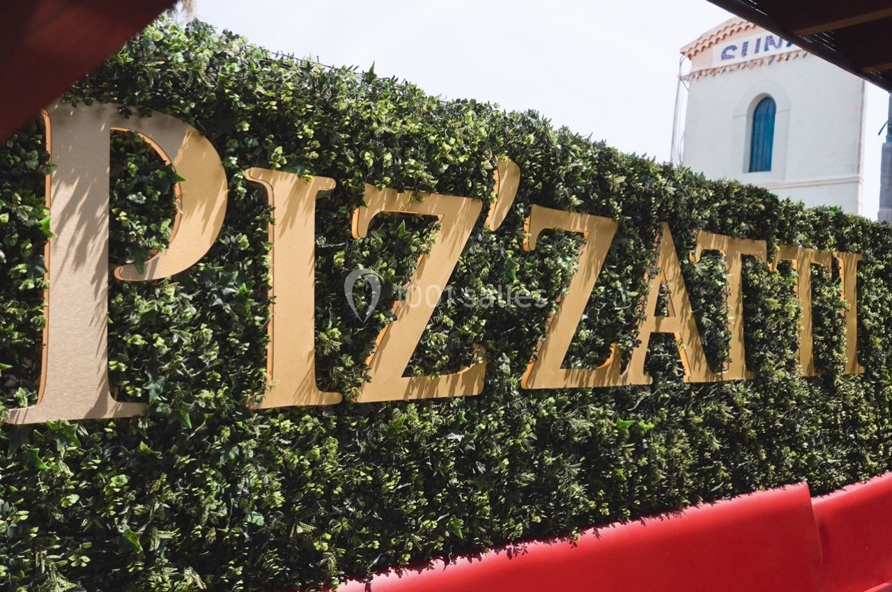 Lettrage doré ’PIZZATTI’ fixé sur une haie de buis artificielle, avec un bâtiment blanc en arrière-plan.