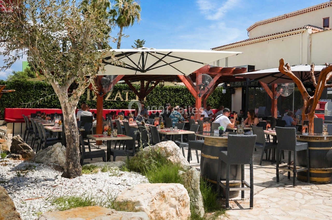 Terrasse extérieure d'un restaurant avec tables, chaises, parasols et clients profitant d'un repas par temps ensoleillé.