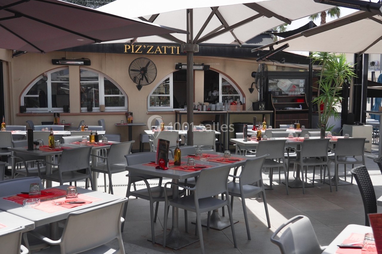 Terrasse d'un restaurant avec des tables dressées, chaises grises et parasols, devant une façade beige avec des fenêtres…