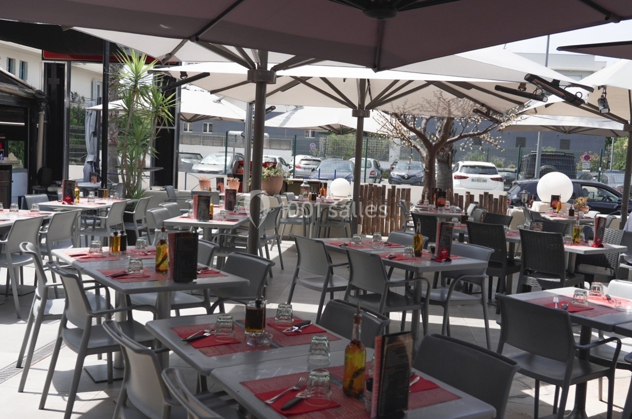 Terrasse de restaurant avec tables dressées, chaises grises, parasols ouverts et vue sur un parking en arrière-plan.