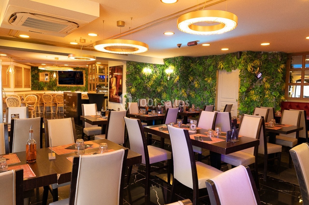 Salle de restaurant moderne avec tables dressées, chaises blanches et mur végétal en arrière-plan.
