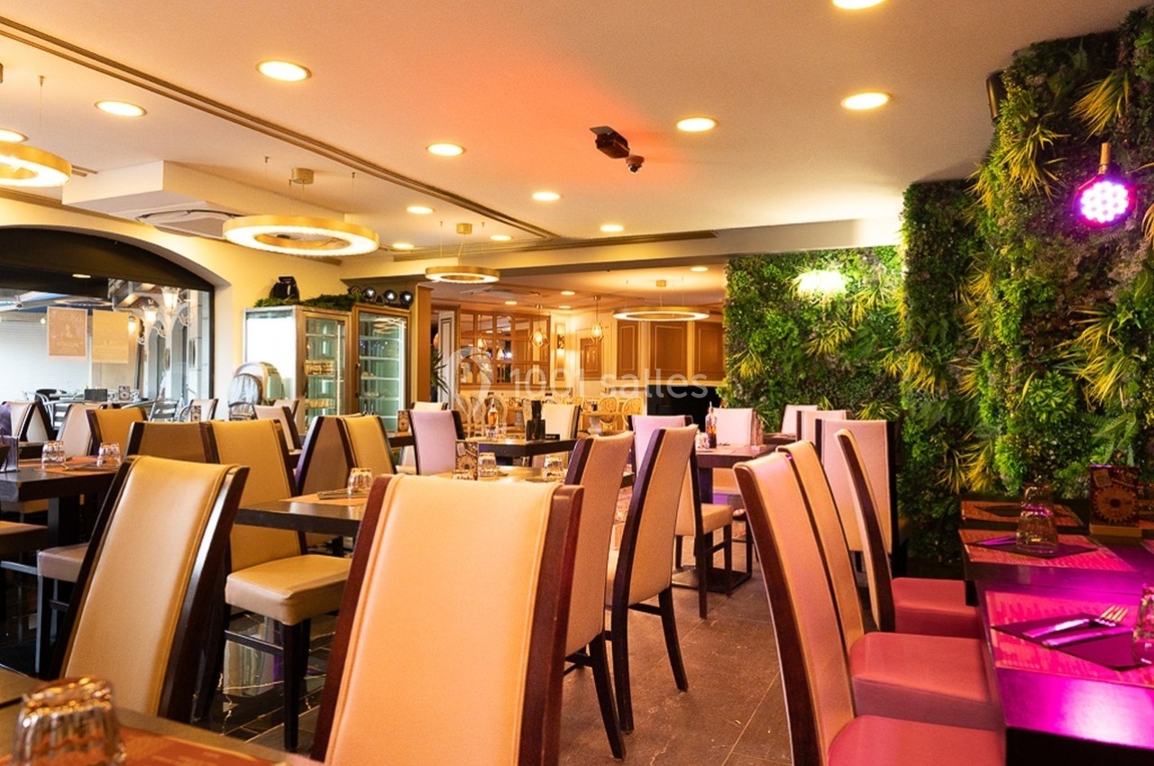 Salle de restaurant lumineuse avec tables dressées, chaises beige et rose, et mur végétal décoratif.