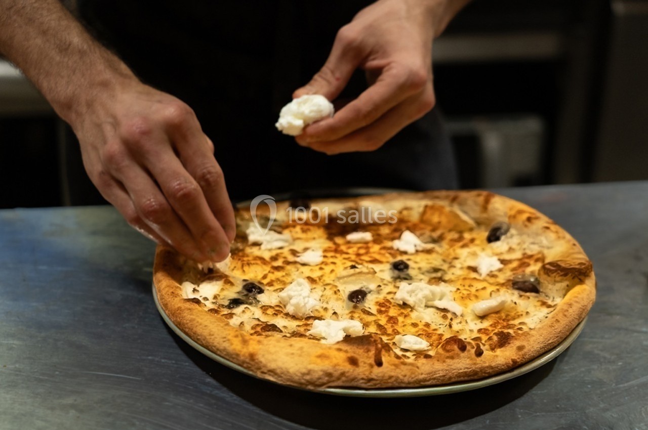 Un cuisinier ajoute des morceaux de fromage sur une pizza garnie d'olives dans une cuisine professionnelle.