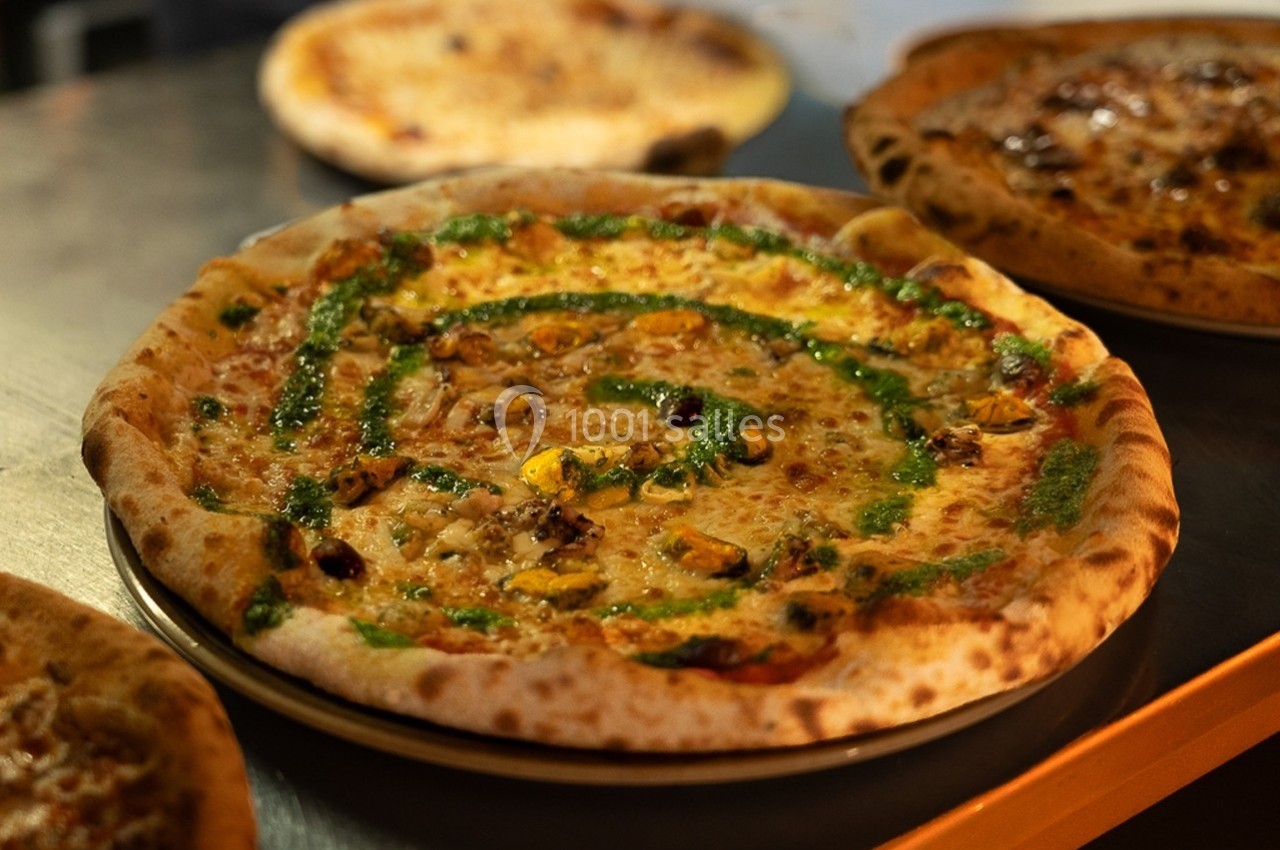 Pizza garnie de légumes, fromage fondu et filet de sauce verte, servie sur une assiette dans un cadre chaleureux.
