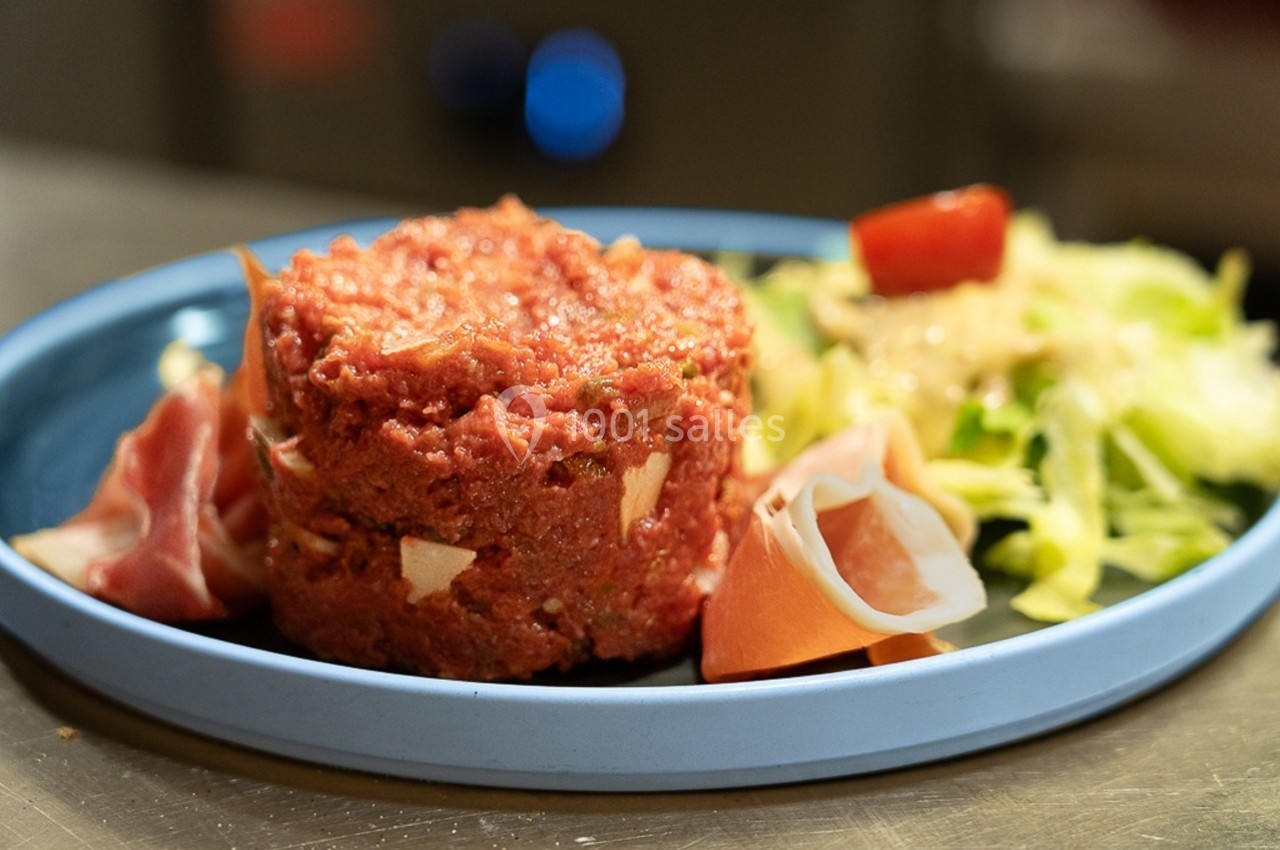Assiette contenant un tartare de viande, des tranches de jambon cru et une salade verte avec une tomate cerise.