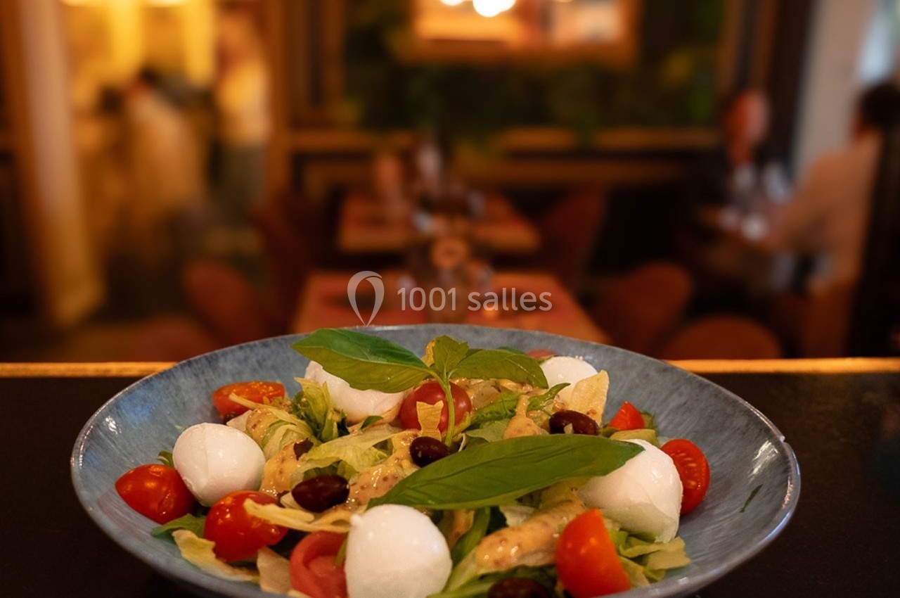 Assiette de salade composée avec tomates cerises, billes de mozzarella, olives et feuilles de basilic, en arrière-plan un…